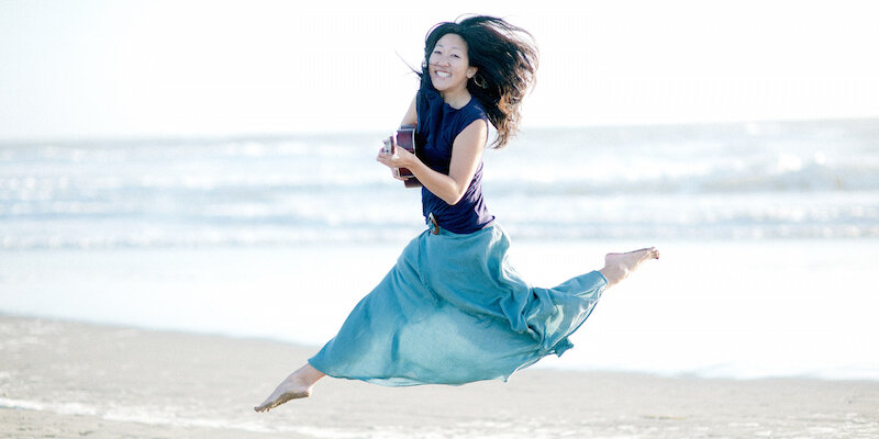 Cynthia Lin smiling, holding an ukulele, leaping in front of a beach